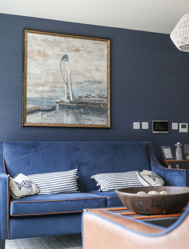 An image of an armchair and blue sofa against a blue wall, with a painting of a harbour on the wall.