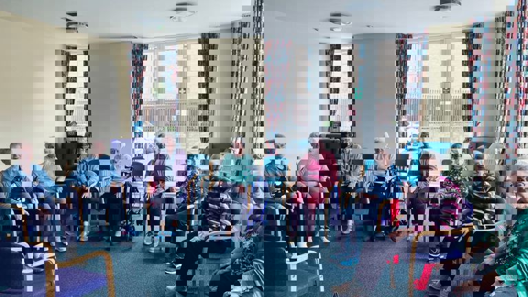 Group of older women sit in chairs in community room